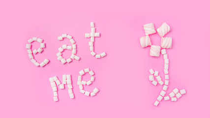 Marshmallows on pink background with sign in English Eat me. Flat lay or top view. Background or texture of colorful mini marshmallows. Winter food background concept.