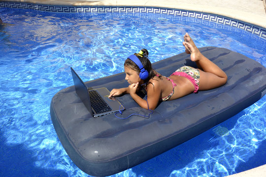 A girl in bikini with computer and headphones sunbathing on an inflatable mattress in the pool