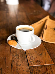 A cup of black coffee with foam in a white mug with a biscuit, stands on a wooden table. Top view with copy space