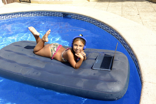 A girl in bikini with computer and headphones sunbathing on an inflatable mattress in the pool