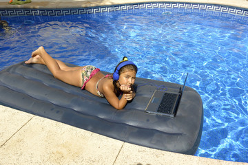 A girl in bikini with computer and headphones sunbathing on an inflatable mattress in the pool