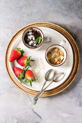 Chocolate and vanilla caramel mug cakes from microwave with fresh strawberries and spoons on round wooden tray over grey texture background. Top view, space