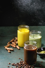 Variety of iced colorful latte drinks. Iced coffee, turmeric and matcha latte cocktails in glass jars with ingredients above over grey green texture background. Copy space