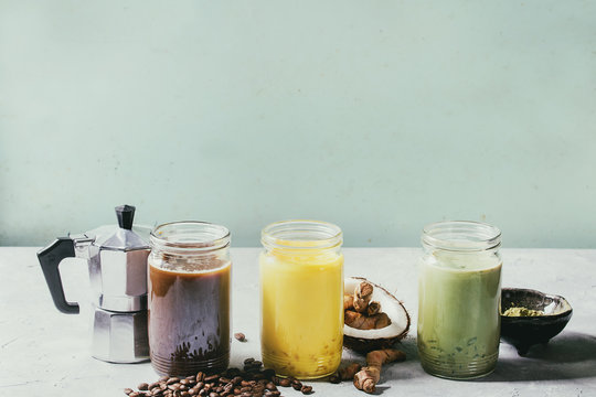 Variety of iced colorful latte drinks. Iced coffee, turmeric and matcha latte cocktails in glass jars with ingredients above over grey green texture background. Copy space. Toned image