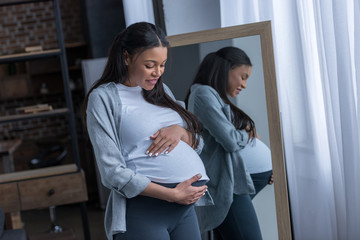 african american pregnant woman looking at her belly while standing at mirror