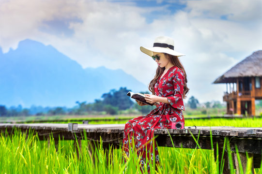 Young woman reading a book and sitting on wooden path with green rice field in Vang Vieng, Laos.