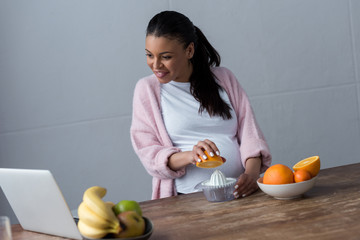 cheerful african american pregnant woman making orange juice with squeezer and looking at laptop