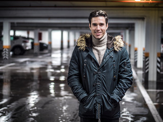 Young trendy man in winter outerwear standing in car parking and smiling to the camera