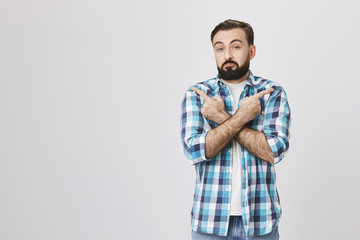 Studio portrait of interesting bearded man with gloomy smile and clueless expression, pointing in different directions with crossed hands, over gray background. Guy do not know which way he should go