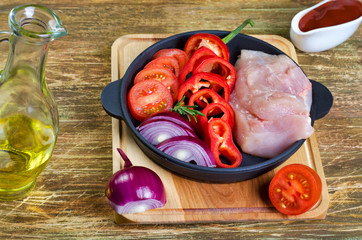 Cooking chicken. Raw chicken breast fillet with red sweet pepper, red onion, tomatoes and rosemary, in the black pan, prepared for cooking. Near to the sauce boat with ketchup and oil in a transparent