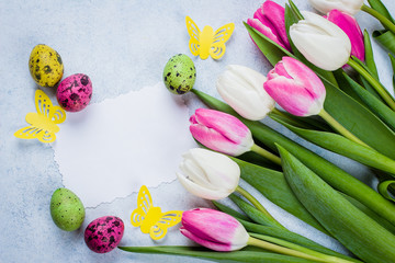 Obraz na płótnie Canvas Spring Greeting card with empty paper blank. Easter Background with white and pink tulips and colorful quail eggs on blue stone table background. Top view, flat lay, copy space.
