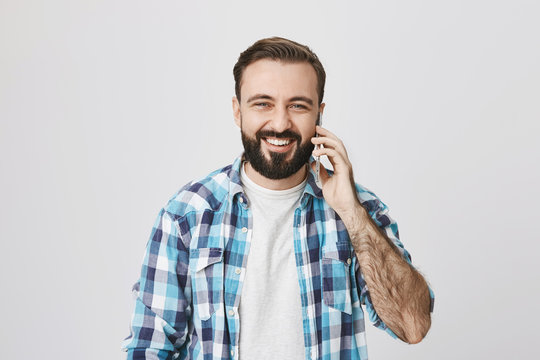 Studio Shot Of Adult Male European With Trendy Haircut And Beard, Smiling Gladly While Talking On Phone And Looking At Camera, Advertising Gadget Over Gray Background. Friends Greet Person With B-day