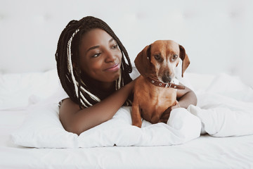 A beautiful African woman lies on her stomach, on a bed with a red dog dachshund, a domestic pet. Good morning. Portrait. Nigeria, Africa.
