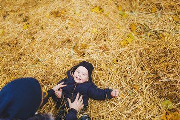 Little cute child baby boy walk in fall park, sit, lay in pile of straw, have fun in dry yellow autumn grass. Mother fun tickle kid son. Parenthood family day 15 of may love parents, children concept.