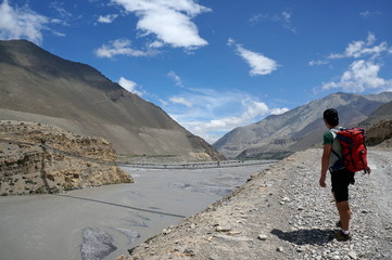 The tourist is standing and looking at the suspension bridge across the Kali Gandaki River, against the backdrop of the Himalayan mountains. Upper Mustang. Nepal. 