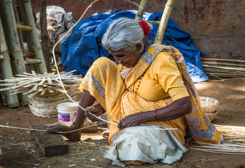 Indian elder lady on yellow saree sitting on the ground making bamboo baskets on the street in...