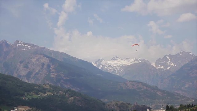 Paragliding in mountain landscape far distance. One paraglider fly using orange parafoil to float descend circling slow low at trees sun snow capped peaks summer video view valley. Air sports tourism