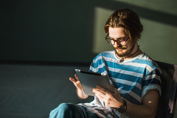 Smiling teenager in glasses using tablet, standing in a dark room near the window, indoor shot in the sunlight
