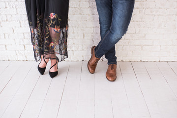 Two pairs of male and female legs in shoes are crossed. Couple standing in front of the white wall crossed-legs in shoes. Girl in black dress with flowers and black shoes. Man in jeans and brown shoes