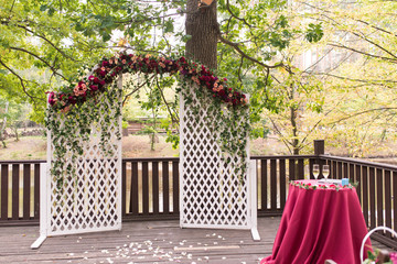 White wooden rustic arch for the wedding ceremony, decorated with red flowers and greenery, is in a...