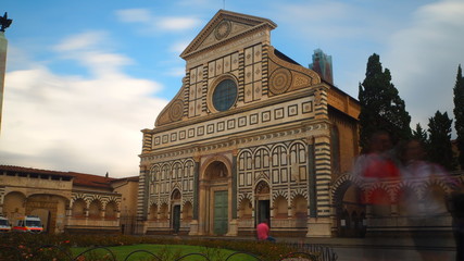 Long exposure of the main facade of the Church of Santa Maria Novella in Florence. The sky has some clouds and there are many tourists in the square in front of it.