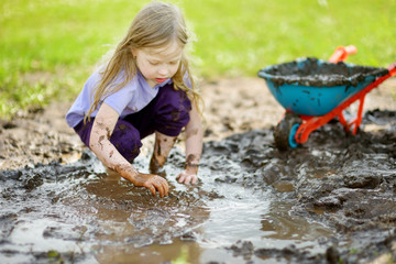 Funny little girl playing in a large wet mud puddle on sunny summer day. Child getting dirty while digging in muddy soil. - obrazy, fototapety, plakaty