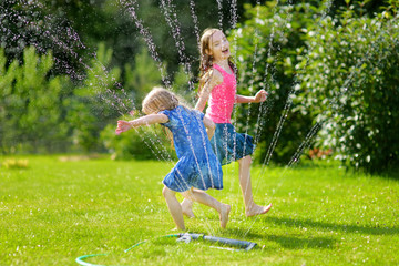 Adorable little girls playing with a sprinkler in a backyard on sunny summer day. Cute children having fun with water outdoors. - obrazy, fototapety, plakaty