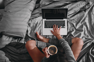 Aerial view of girl sitting on the bed and using laptop while sitting and driknking coffee in the morning