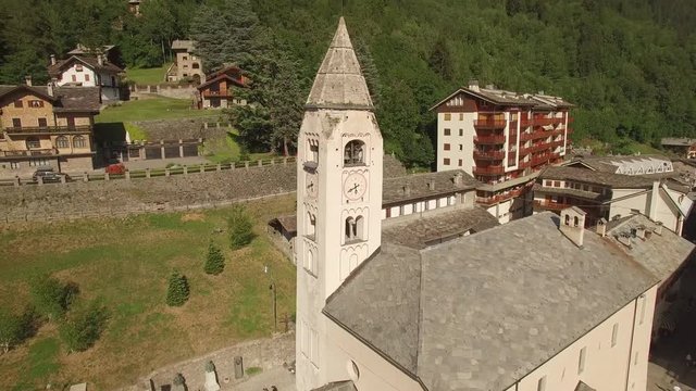 Aerial shot of old belfry with bell ringing alarm over medieval European town in Alps valley Italy. 4k copter close flight around St. Pantaleon Church clock tower. Medieval history historic heritage