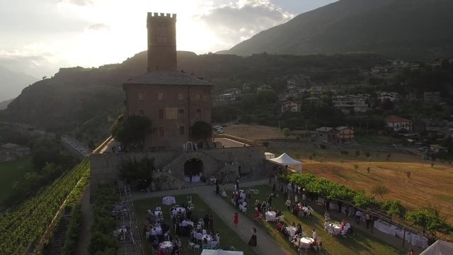 4k Aerial shot of open air gathering people celebrating wedding event by old castle vertical pan tracking. Sun shine over medieval chateau vineyard and fields in Alps mountains scenic view from above