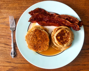 Breakfast of stacks of pancakes, syrup, bacon strips, set up on a plate with a fork.  No people.