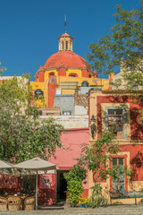 A partial view of an outdoor restaurant with umbrellas, an iron bench, greenery, colorful buildings and part of a church with a dome top, in the San Fernando Plaza, in Guanajuato, Mexico - 191672418