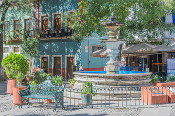 Fountain surrounded by a green iron fence, a green iron bench, some greenery, umbrellas for dining, and colorful two-story buildings, in the San Fernando Plaza, in Guanajuato, Mexico