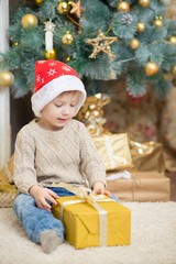 a boy in a red hat sits at a Christmas tree with a gift