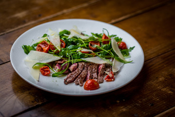 top view of italian sliced beef salad with rucola, tomatoes and parmesan cheese on white plate on wooden table