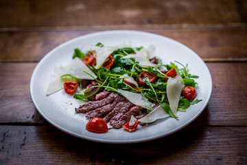 top view of italian sliced beef salad with rucola, tomatoes and parmesan cheese on white plate on wooden table