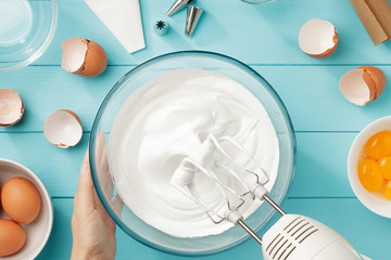 Whipped egg whites cream to perfect peaks in glass bowl with mixer and hand on blue wooden table. - 191681440