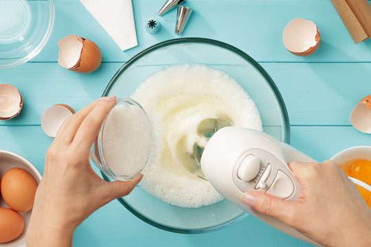 Female hands add a sugar to the bowl with whipping egg whites with mixer.