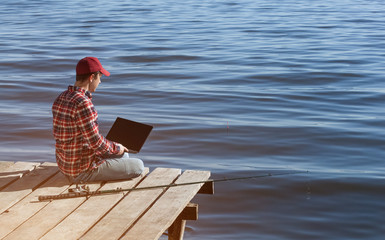 A fisherman man works on a laptop, sits on a wooden pier near the lake, next to it there is a fishing pole. © Dimid