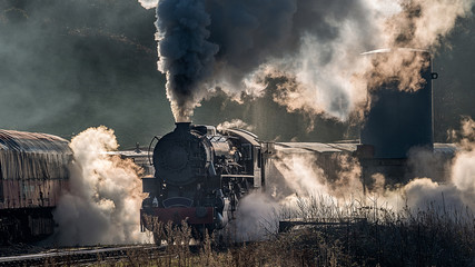 Steam train locomotive approaching a station passing through a goods yard letting of smoke and steam lit from behind creating atmospheric photograph