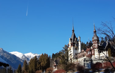 Landscape with snow-capped mountains, Peles palace, blue sky and white  track of plane.