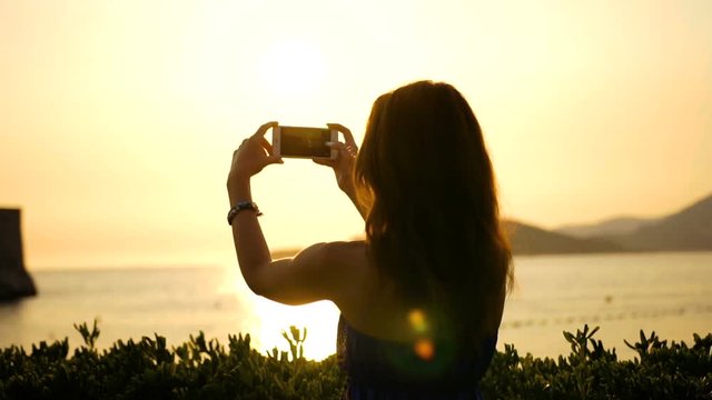 Back view on the young woman taking photos on her smart phone of the beautiful sunset over sea on the coastline with mountains far on the horizon. Outside