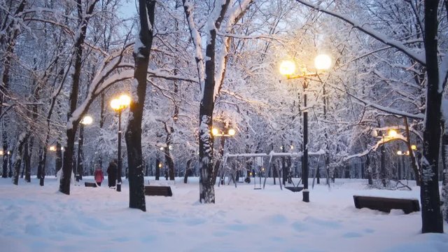 Beautiful evening park winter scene, trees and ground are covered with snow, yellow street lights shining, people are walking on background, russian Moscow park.