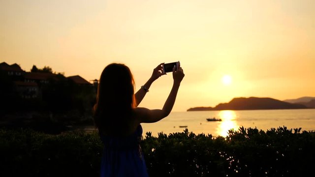 Rear of the silhouette of young woman taking a photos on the smart phone of the beautiful sunset over sea on the coastline with mountains far on the horizon. Outside