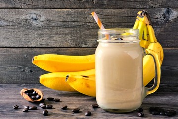 Coffee, banana smoothie in a mason jar with coffee beans and bananas in background. Side view against a rustic wood background.
