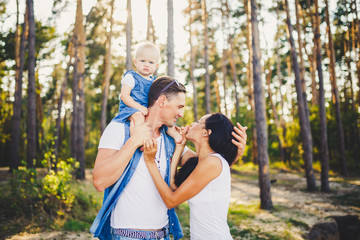 Fototapeta premium Family mom, dad and daughter sits at the daddy on the shoulders, and parents kiss on the nature in the forest in the summer at sunset