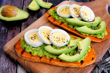 Sweet potato toasts with avocado, eggs and chia seeds on a wood board. Table scene with a wooden background.