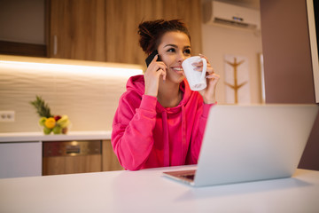 Relaxed smiling young brunette woman drinking coffee or tea, talking on mobile and using laptop.