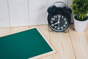 Blank green chalkborad with wood frame and little decorative tree in white vase and vintage alarm clock on wooden table with copy space.