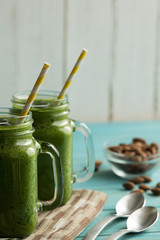 Two green healthy smoothies in mason jar mugs with straws on a table with nuts